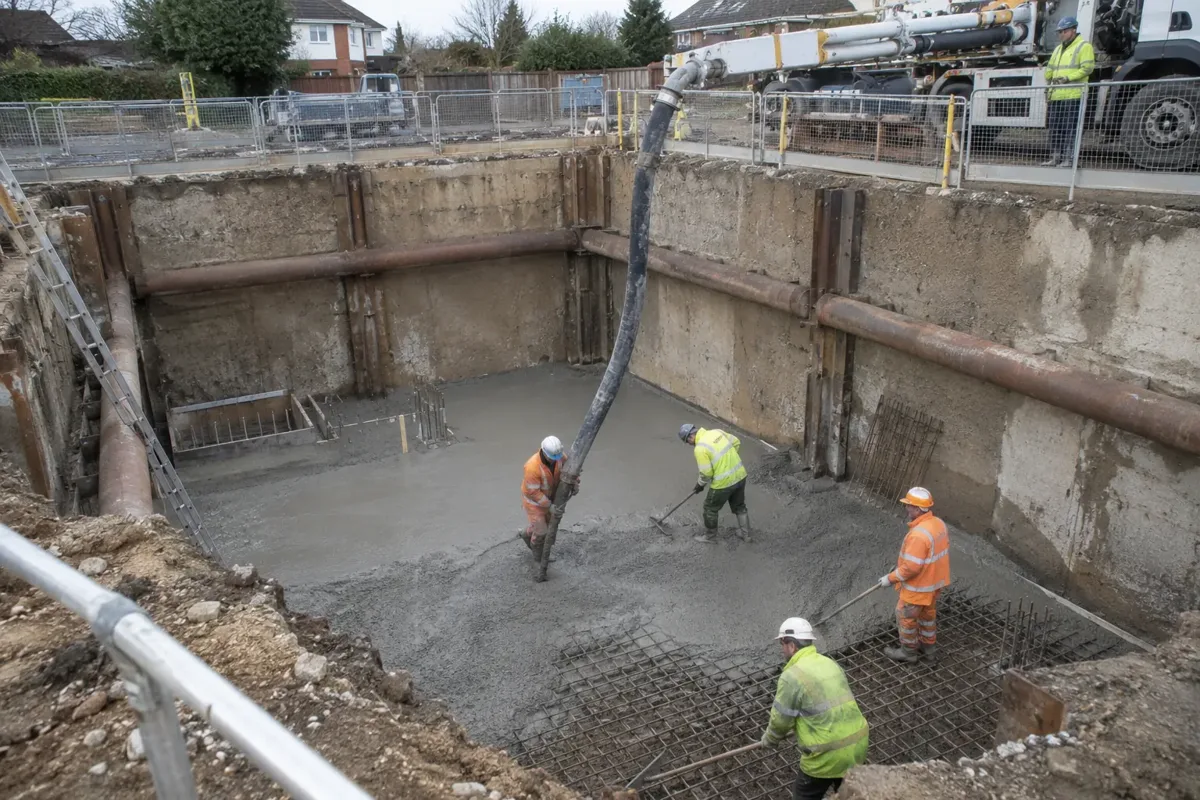 Wide view down into an open, safely shored basement excavation where a thick concrete delivery hose descends from ground level to a PPE-clad crew placing concrete onto a prepared basement floor. Guarded excavation rim, retaining supports and clear below-ground depth.
