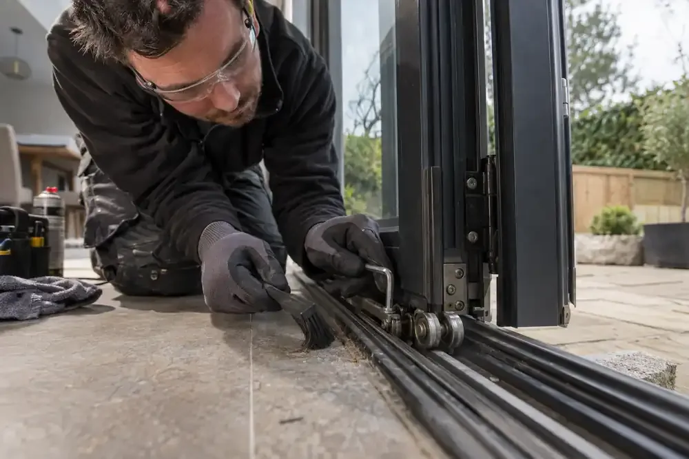 Macro low-angle trade photograph: kneeling technician servicing the bottom roller carriage and cleaning the track of partially folded patio bifold doors, small brush and hex key, glass visible above.